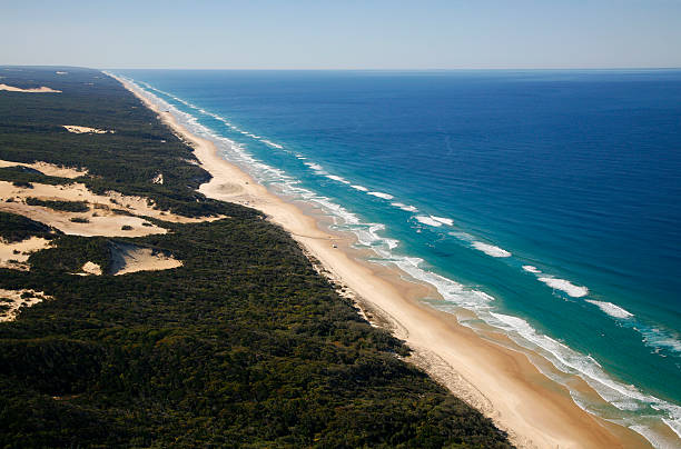 Aerial view of spectacular Fraser Island coastline.  The long sandy beach stretches between sand dune covered rain forest and aqua blue ocean. Lines of surf crash onto beach.  A few vehicles can be seen on the beach.