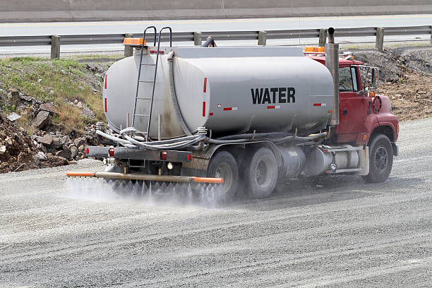 Water truck sprays water on newly graded gravel prior to being compacted with a steam roller on a new road construction site.