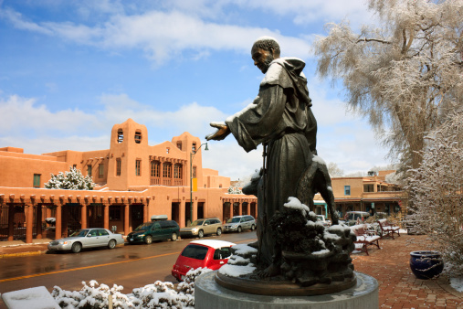This is a picturesque view of the historic Downtown Plaza in Santa Fe New Mexico. A light snow covers the statue of St. Francis of Assisi, brought to the grounds of the St. Francis of Assisi Cathedral and Basilica in 1967. The statue represents the patron saint of the Catholic Diocese of Santa Fe, illustrating the Catholic Religious heritage of this region.