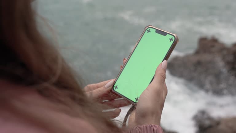 Woman Holds a Phone with a Green Screen and Tracking Markers, Standing on a High Cliff with Waves Crashing Against the Rocks in the Background.