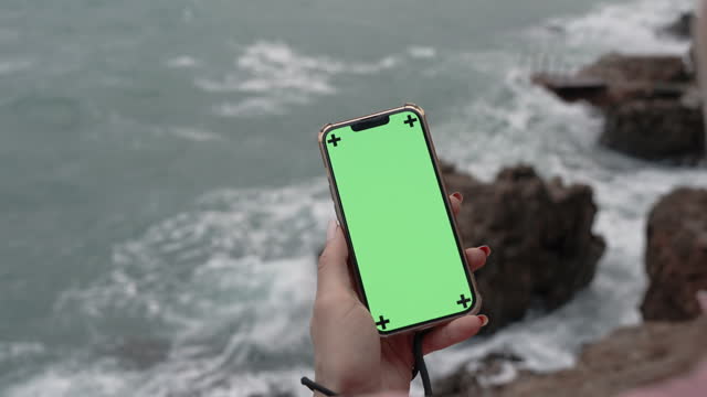 View From Behind the Female Head as She Holds a Smartphone with a Green Screen While Standing on the Edge of a Cliff by the Stormy Sea with Large Waves and Overcast Weather.