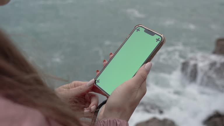 Smartphone with a Green Screen and Tracking Markers is Held by a Woman with Long Hair Standing on the Edge of a Cliff by the Sea. Overcast Weather and Large Waves.