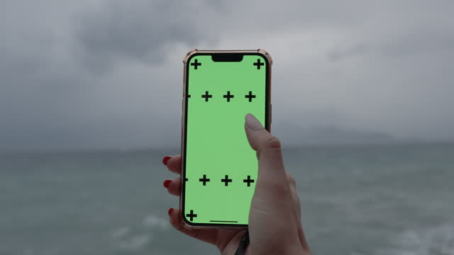 Smartphone with a Green Screen and Tracking Markers in a woman's hand, swiping sideways, against the backdrop of cloudy and rainy weather at the sea.