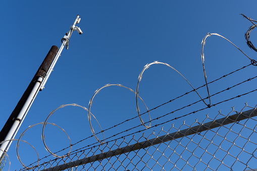 Fence with razor wire with blue sky in background. \nRazor wire is made up of high tensile core wire and a punched steel tape with sharp barbs at close intervals uniformly.