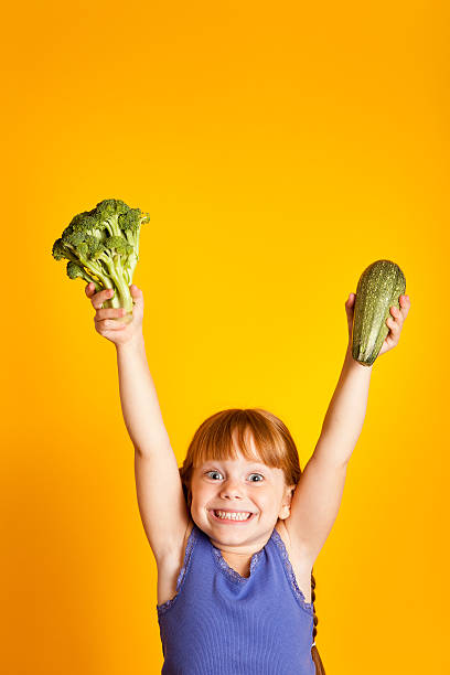 aufgeregt und lächelnd mädchen halten, brokkoli und zucchini - child jumping vegetable food stock-fotos und bilder