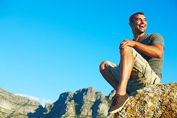 istock Happy young man sitting on rock against blue sky 182880879