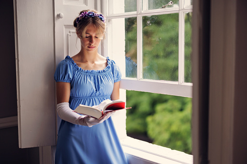 Young woman wearing a regency era dress is reading a book near a window.\nShot with Canon R5