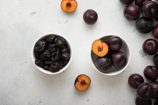 Two bowls with dried prunes and ripe raw plums on light background.
