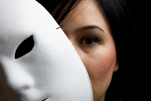 A young woman with black hair and black eyes looking at the camera from behind a white mask.The photo was shot with a full from DSLR camera in horizontal close up composition.The mask is on the left side of frame. 