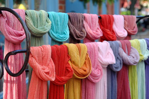Many colored hip scarves on a wrought iron display rack. Photo was taken outside a boutique in a trendy downtown section Portland, Maine. The time is summer in the late afternoon. The picture offers a selection from pale pinks and muted green to bright reds and turquoise.