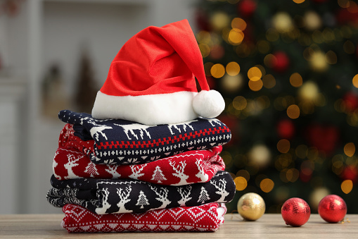 Stack of different Christmas sweaters, Santa Claus hat and decorative balls on table against blurred lights