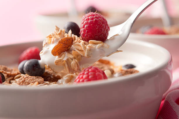 Pouring milk on a bowl of cereal (muesli/granola) with fresh strawberries, blueberries and fruit in the background.
