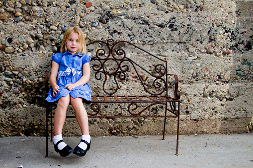 Pretty blond 8 year old girl sitting on a wrought iron bench in front of a concrete wall