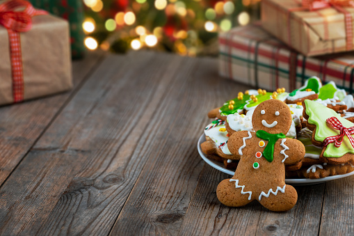 Gingerbread man cookie and plate with gingerbread cookies decorated with icing on wooden table. Defocused lights and Christmas gifts on background. Concept of preparing Christmas gifts for the winter holidays. Copy space.