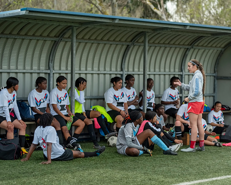 Female soccer coach explaining game plan to girl players sitting on bench on soccer field