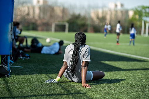 Rear view of a young female soccer reserve goalie sitting on sideline and watching a game on the field