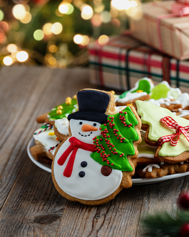 Christmas greeting card with gingerbread cookie in the shape of snowman with Christmas tree and plate with gingerbread cookies decorated with icing on wooden table. Defocused lights and Christmas gifts on background.