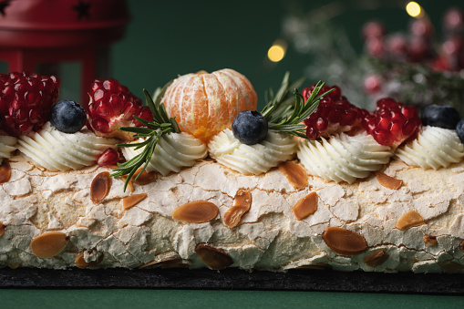 Meringue roll sprinkled with powdered sugar decorated with blueberries, pomegranate and almond flakes on the dark green background with blurred Christmas tree. Christmas festive pastry