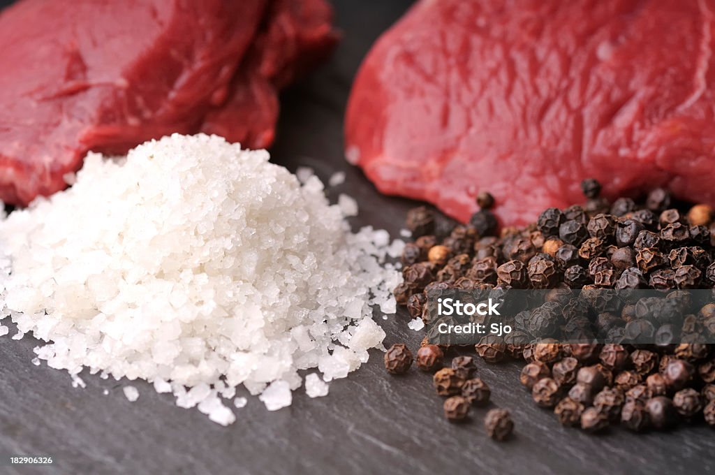 Steaks  A culinary themed, macro photograph of two raw steaks, a pile of kosher sea salt, and a pile of whole peppercorns. The ingredients are arranged on top of a dark grey stone surface. The shot primarily focuses on the sea salt and peppercorns, while the two cuts of steak are out of focus in the background.  Animal Blood Stock Photo