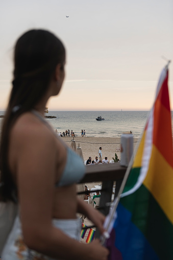 Tel Aviv, Israel - June 8, 2023: Late on a summer afternoon, a police boat is seen near the beach in Tel Aviv. In the foreground stands a young woman holding a rainbow flag.