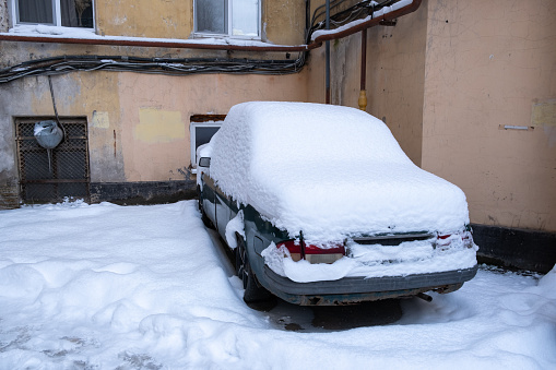 An abandoned car covered in snow in the backyard of an old house after a snowfall