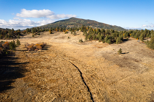 Captured from above, this aerial view showcases the rich tapestry of Julian, Stonewall Mine Trail, in Cuyamaca Rancho State Park, San Diego County, California. Evergreen and autumn-kissed trees create a vibrant mosaic around a serene clearing, nestled between majestic mountains, all under the canvas of a blue cloudy sky
