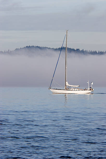 Sailboat in Morning Fog "Sailboat motors along in a quiet bay with a fog bank approaching and mountains visible beyond. Taken on Puget Sound, Washington State." bainbridge island photos stock pictures, royalty-free photos & images