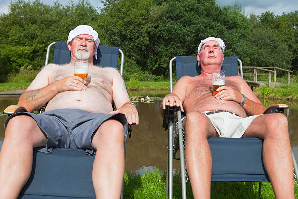 Two Old Men Sunbathing Two older overweight men with handkerchiefs on their heads to protect them from sunburn enjoy a picnic with just a glass of beer out in the countryside. hairy fat man pictures stock pictures, royalty-free photos & images
