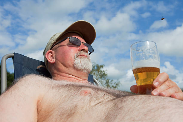 Obese Man With A Glass Of Beer Low viewpoint shot of a 60s man enjoying the Summer sun with a glass of beer resting on his enormous pot belly. hairy fat man pictures stock pictures, royalty-free photos & images