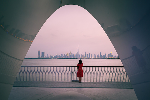 Woman standing enchanted, gazing at the breathtaking Dubai skyline from Creek Harbour. The city comes alive with twinkling lights against the dusky sky, creating a scene of tranquil beauty and urban allure