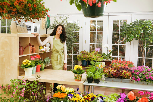 A small business owner of a retail flower shop. A black African American woman florist entrepreneur shopkeeper. She is working close to the checkout counter in her shop, looking at the camera posing with various colorful potted plants and flower display in the background. Photographed in horizontal format.