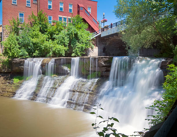 Water cascading over Chagrin Falls and into the Chagrin River  in the village of Chagrin Falls, Ohio. The red brick building with its stone foundation and the lush green folliage help to accent the brillian blue sky. A lamp post with hanging baskets adorn the bridge over the waterfall.