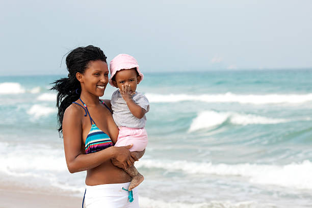 Young woman holding baby girl, standing on coastline of sea.