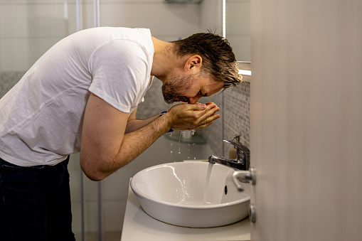 The mid adult man is seen using a facial scrub to exfoliate his skin while washing his face in the bathroom sink, preparing for the day ahead