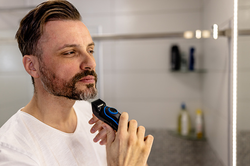A mid adult Caucasian man is carefully using an electric razor to shave his beard in the bathroom. With precision and ease, he expertly removes the hair to reveal a smooth and clean-shaven face