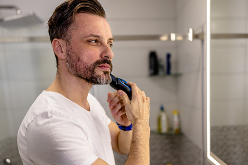 In the bathroom, a mid adult Caucasian man is using an electric razor to shave his beard. The ease and convenience of the razor make the process quick and effortless, allowing him to look his best with minimal effort