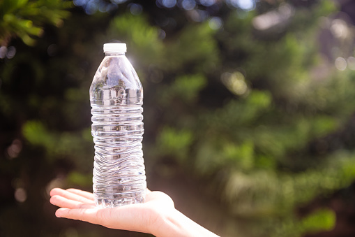 This is a photograph taken outdoors on a sunny day of a plastic water bottle held by an unrecoginezable Hispanic Woman in her 20s with a green background.