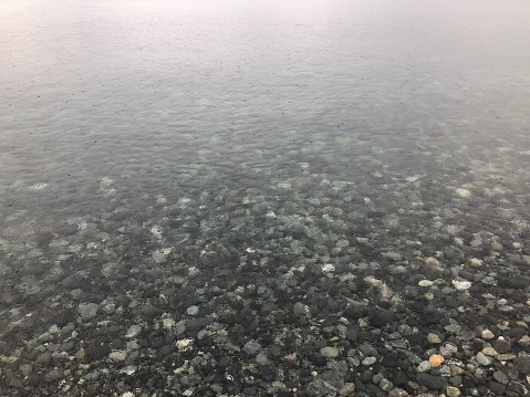 Clear water shows the stones on the ocean bed below, while raindrops create patterns on the surface of the water.