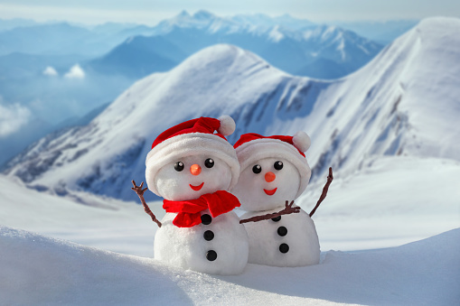 View of a couple of snowmen in a santa hats on their heads against the backdrop of snowy mountains. Holiday christmas background with copy space for text