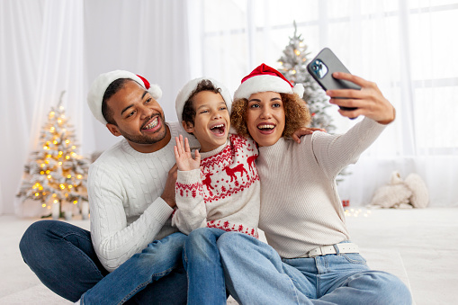 African-American family celebrates Christmas with their child and takes selfie on smartphone at home, boy with his parents in Santa hat takes pictures online and waves his hand in greeting
