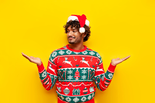 young indian man in new year clothes holds empty hands at sides and chooses on yellow isolated background, pensive hindu guy in santa hat and christmas sweater advertising copy space