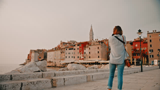 SLO MO Rear view of female tourist photographing old town while exploring Rovinj from pier by sea in Croatia