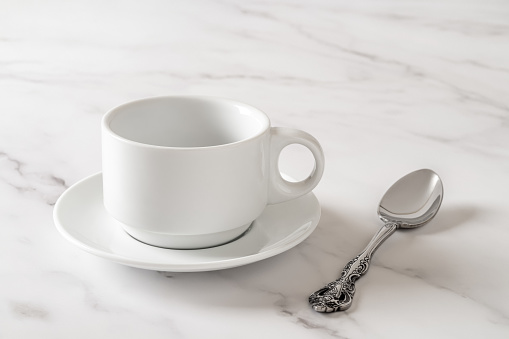 White cup on saucer and teaspoon over marble table. Empty clean porcelain crockery for drink design mockup. Tableware, breakfast, tea and coffee concept. High key image. Front view.