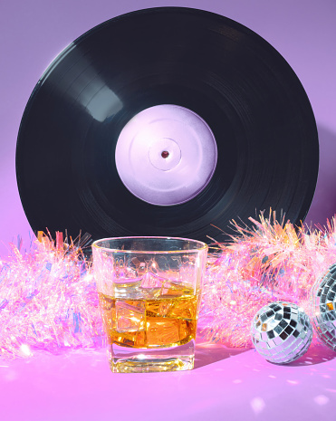A Glass of Scotch Whiskey, A Vinyl Record , Mirror Ball Disco Balls and Holographic Holiday Christmas Tinsel on a Pastel Pink and Purple Background