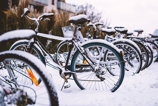 The snow covered bicycles are parked in a bike rack. The bicycles are old-fashioned with large fenders and a basket on the front.