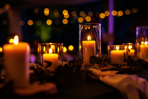 The table is decorated with lit candles in glass vases and greenery, creating a warm and romantic atmosphere. The tablecloth is white, and there are folded napkins on the table.
