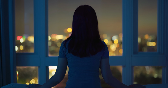 Rear view of woman sitting alone on bed in room and looking through window at night