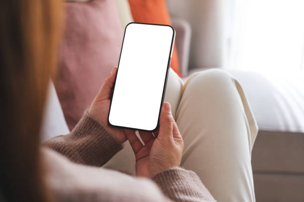 istock Mockup image of a woman holding mobile phone with blank desktop white screen while sitting on a sofa at home 1829571892