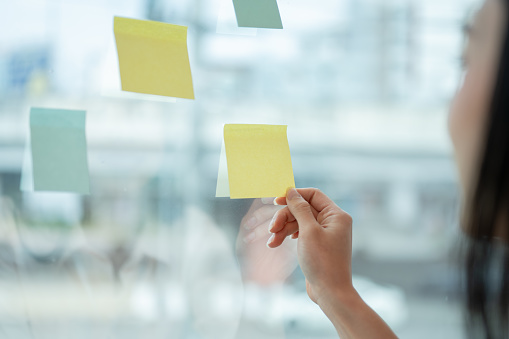 Close-up image of a businesswoman or female marketing officer attaching or looking at a sticky note on a glass wall. An empty sticky note mockup. ideas, memo, brainstorming