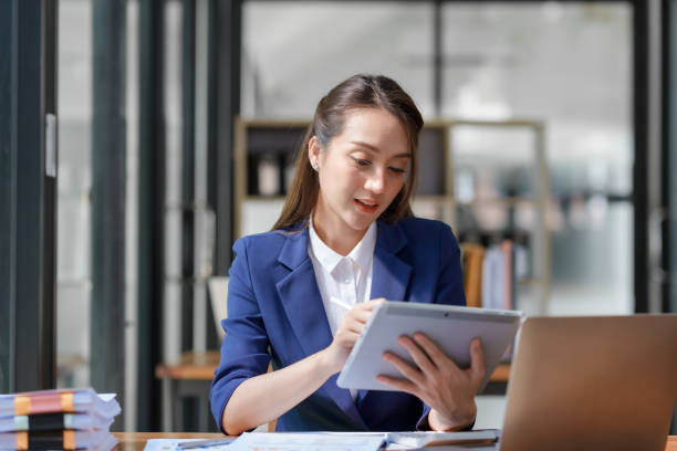 istock Asian business woman sitting happily with laptop and using tablet and taking intense notes on the work clipboard and smiling happily on the assignment. 1829694924